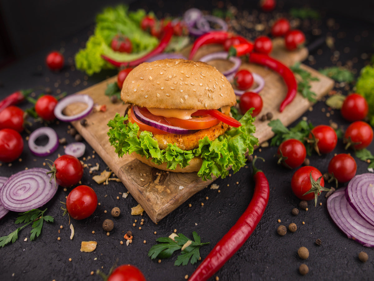 delicious looking chicken burger served on a wooden platter surrounded by vegetables such as onion, tomatoes and chilli